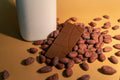 Milk chocolate bar sitting atop roasted cocoa beans and a glass jar of milk