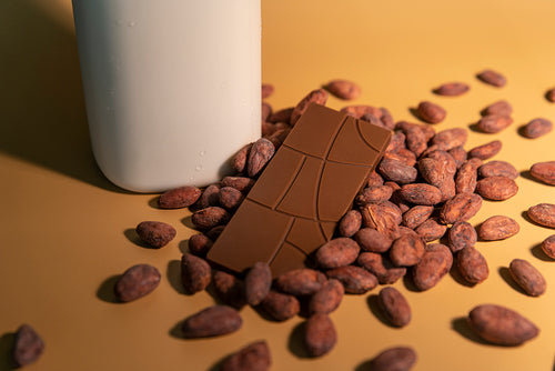 Milk chocolate bar sitting atop roasted cocoa beans and a glass jar of milk